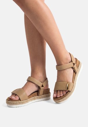 Piernas que llevan sandalias de cuero beige con tiras en el tobillo, suelas gruesas tejidas y adornos metálicos dorados decorativos en las tiras delanteras.