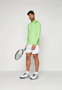 Camisa de manga longa verde-lima, calções brancos e ténis brancos com detalhes em azul-marinho. A segurar uma raquete de ténis com um punho azul e preto.