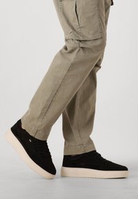 Zwarte suede sneakers met een dikke witte zool, met een gladde afwerking, vetersluiting en subtiele branding aan de zijkant. Gerd gedragen met lichtbeige broeken.
