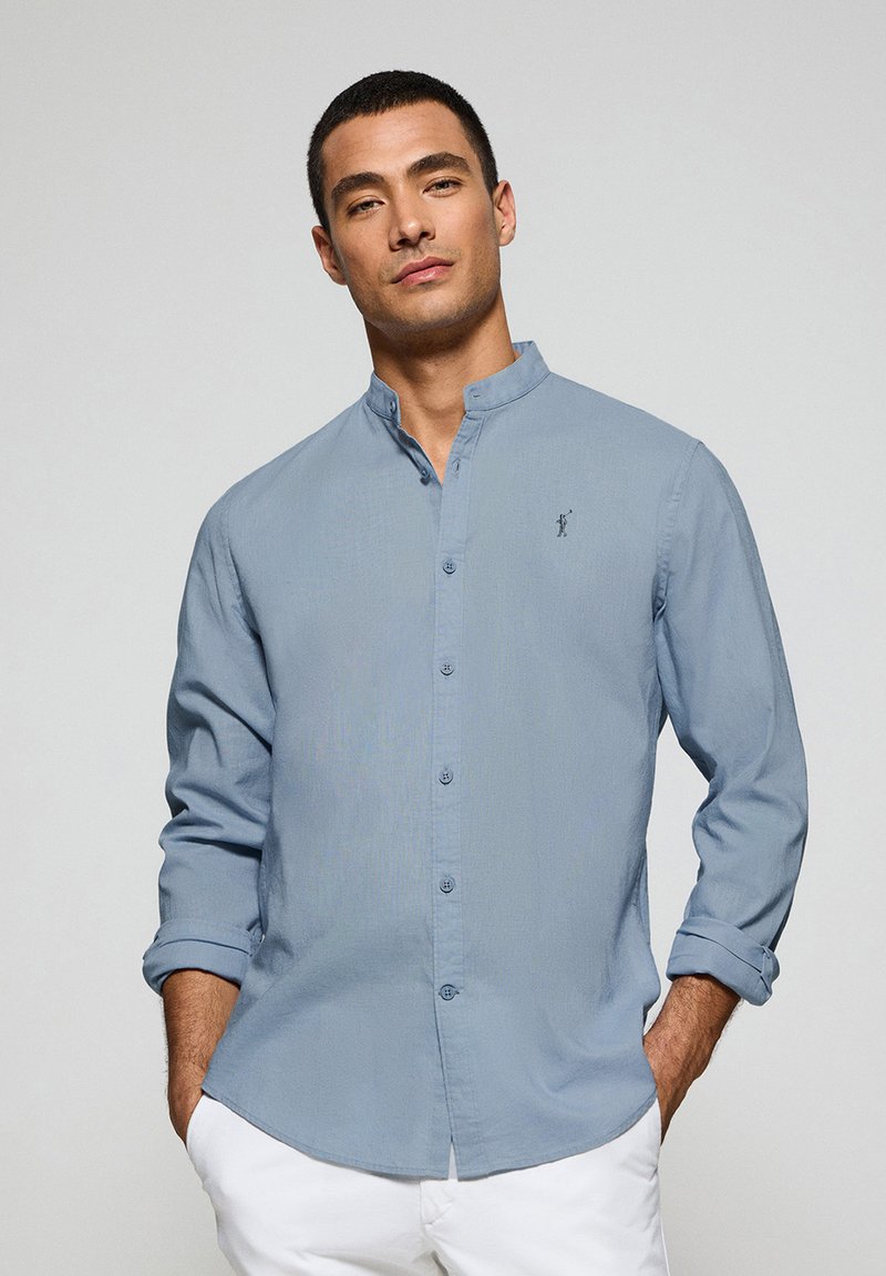 Ljusblå knapp-up skjorta med bandkrage. Har långa ärmar och en liten broderad logotyp på bröstet. Slät tygtextur.