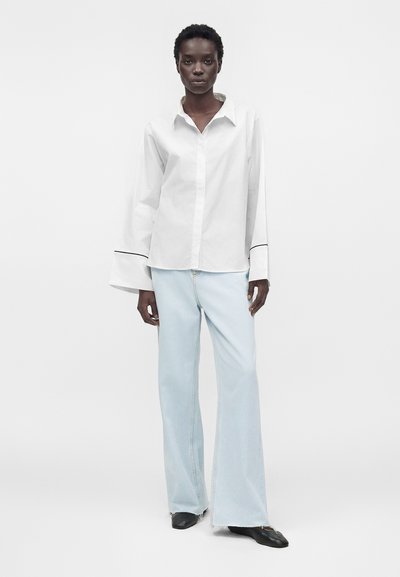 Personne portant une chemise blanche à manches longues avec de larges poignets et un jean bleu clair à jambes larges, debout devant un fond blanc uni.