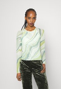 T-shirt de manga longa com um padrão em mármore verde, branco e azul claro; tecido leve e texturizado; combinado com calças de veludo verde escuro.