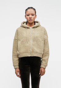 Μπεζ ψεύτικη γούνα cropped hoodie με φερμουάρ μπροστά, κορδόνι και πλούσια υφή. Φοριέται με εφαρμοστούς σκούρους καφέ παντελόνες.