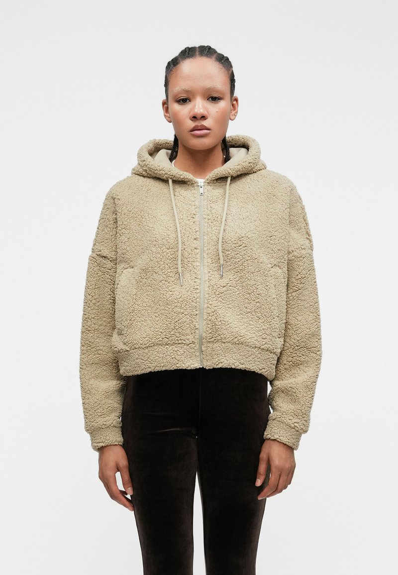 Μπεζ ψεύτικη γούνα cropped hoodie με φερμουάρ μπροστά, κορδόνι και πλούσια υφή. Φοριέται με εφαρμοστούς σκούρους καφέ παντελόνες.