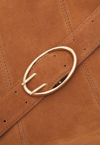 Zamszowy pasek w ciepłym brązowym kolorze, z dużą złotą okrągłą klamrą i przeszyciami na pasku. Widoczne otwory do regulacji.