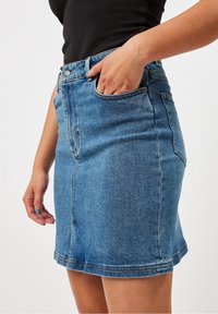 Femme portant une jupe en denim bleu et un haut noir à côtes, avec la main droite reposant à l'intérieur de la poche de la jupe, sur fond uni.