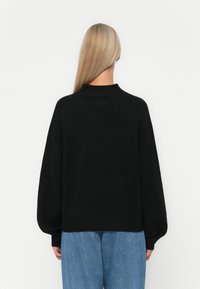 Svart strikket genser med høy krage, vide ermer og ribbestrikket kant. Båret over lyseblå denim, har en teksturert stoff.