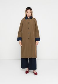 Woman standing against white wall wearing brown coat with navy collar and cuffs, dark wide-leg pants, and red pointed flats.