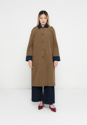 Donna in piedi contro un muro bianco, indossa un cappotto marrone con colletto e polsini blu navy, pantaloni larghi scuri e ballerine rosse a punta.