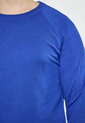 Close-up van een persoon die een helderblauwe ronde hals trui draagt met zichtbare stiksels op de schouder en de halslijn.
