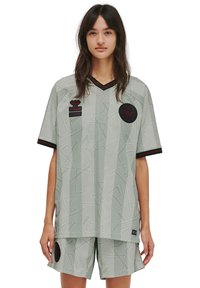 Ung kvinna iklädd en ljusgrå mönstrad sporttröja och shorts med svarta och röda laglogotyper och detaljer, stående mot en vit bakgrund.