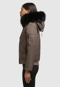 Braune gepolsterte Jacke mit einer schwarzen Fellkapuze. Verfügt über eine Reißverschlusstasche am Ärmel und seitliche Taschen. Gerippte Bündchen und Saum runden das Design ab.