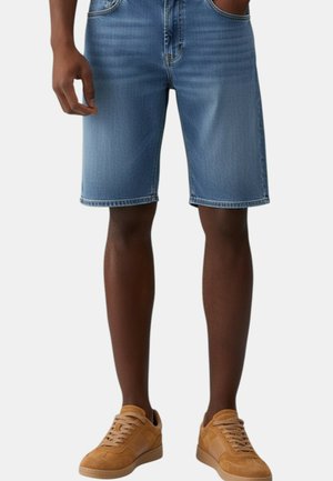 Uomo con pantaloncini di jeans blu medio e sneakers in pelle scamosciata color cuoio, in piedi con una mano in tasca su uno sfondo semplice.