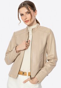 Beige leren jacket met een hoge kraag, ritssluiting en gestikte details. Soepele textuur en getailleerd ontwerp. Draagt over een lichte top.