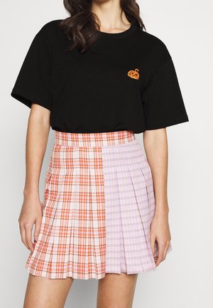 Kobieta w czarnej koszulce z małą grafiką pomarańczowej dyni oraz plisowanej mini spódniczce w kratkę z pomarańczowymi i fioletowymi wzorami.