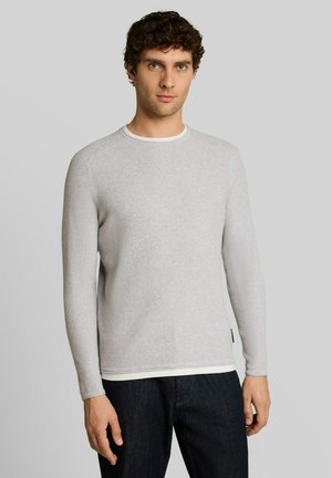 Mann mit kurzen lockigen Haaren, der einen hellgrauen, langärmeligen Strickpullover und dunkle Jeans trägt, vor einem schlichten grauen Hintergrund steht.