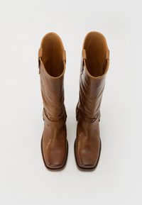 Bottes en cuir marron jusqu'au genou avec un bout carré, une texture lisse et une sangle décorative autour de la cheville.