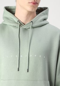 Světlězelená mikina s kapucí na stahovací šňůrku, vybavená přední kapsou ve stylu klokánka a bílým nápisem "JACK & JONES" přes hruď.