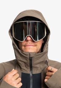 Chaqueta impermeable marrón con un alto cuello, detalles en negro y cierre de cremallera. Modelo luciendo gafas de esquí espejadas grandes, mostrando un diseño elegante.