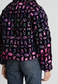 Gevoerde jas in zwart, paars en roze met een geometrisch patroon. Voorzien van een hoge kraag en elastische manchetten, gecombineerd met blauwe jeans.