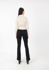 Cremfarbener Rippstrickpullover mit Kragen, kombiniert mit schwarzen Schlaghosen und schwarzen Stiefeletten mit Absatz, von hinten vor einem weißen Hintergrund gezeigt.