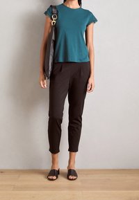 Donna che indossa una maglietta verde acqua a maniche corte, pantaloni neri a vita alta, ciabatte aperte nere, mentre tiene una borsa a spalla nera con dettagli dorati, in piedi su un pavimento di legno.
