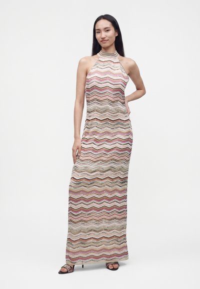 Femme portant une robe longue sans manches à col montant avec un motif zigzag multicolore, debout avec une main sur la hanche sur un fond uni.
