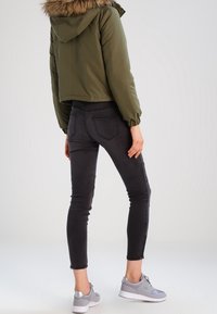 Olivově zelená zkrácená bunda s kapucí podšitou kožešinou, spárovaná s černými skinny džíny a šedými sportovními botami. Hladké materiály a přiléhavý design.