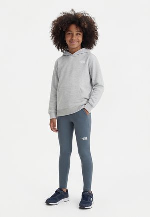 Kind mit lockigem Haar trägt einen hellgrauen Kapuzenpullover, dunkelgraue Leggings und marineblaue Turnschuhe, steht mit einer Hand in der Tasche und lächelt.