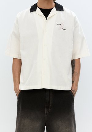 Personne portant une chemise crème à manches courtes avec un col noir et le texte "GOOD PLACE" sur la poitrine, associée à un jean noir délavé et des bracelets au poignet.