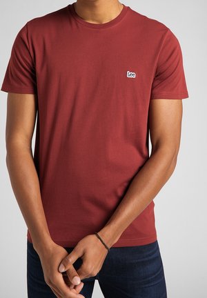 Uomo che indossa una maglietta Lee bordeaux a maniche corte e jeans scuri, con le mani incrociate davanti, su uno sfondo chiaro e uniforme.
