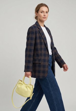 Vrouw met een marineblauwe geruite blazer, wit overhemd en blauwe spijkerbroek, die een kleine gele handtas vasthoudt en loopt tegen een effen lichte achtergrond.