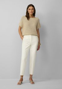 Beige gebreide top met korte mouwen, gecombineerd met witte op maat gemaakte broeken. Model draagt open-tanden sandalen met dunne banden en een gouden armband.