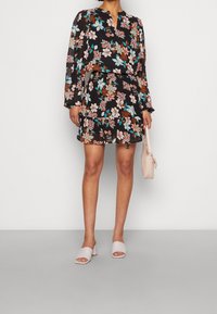 Blomsterkjole med sort baggrund, lange gennemsigtige ærmer og flæset kant. Et par lyserøde slip-on sko og en matchende håndtaske.