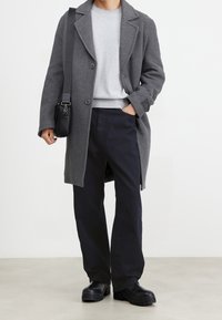 Grå yllekappa med snedskurna slag, grå tröja, mörka byxor och svarta grova stövlar. Svart budväska hänger från vänster axel.