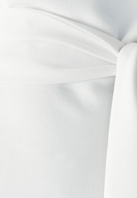 Bijela tkanina s mekanim, teksturiranim izgledom, s dizajnom koji se omata i detaljem s vezom na struku.