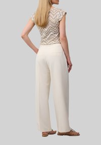 Beige Oberteil mit Zickzackmuster und kurzen Ärmeln, kombiniert mit weißen weitgeschnittenen Hosen. Leichter Stoff, minimale Nähte und sichtbare Gesäßtaschen.