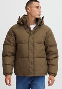 Smeđa puffer jakna s visokim ovratnikom, koja ima kvadratni dizajn, prednje kopčanje na snap i elastične manžete. Nošena preko bijele košulje.