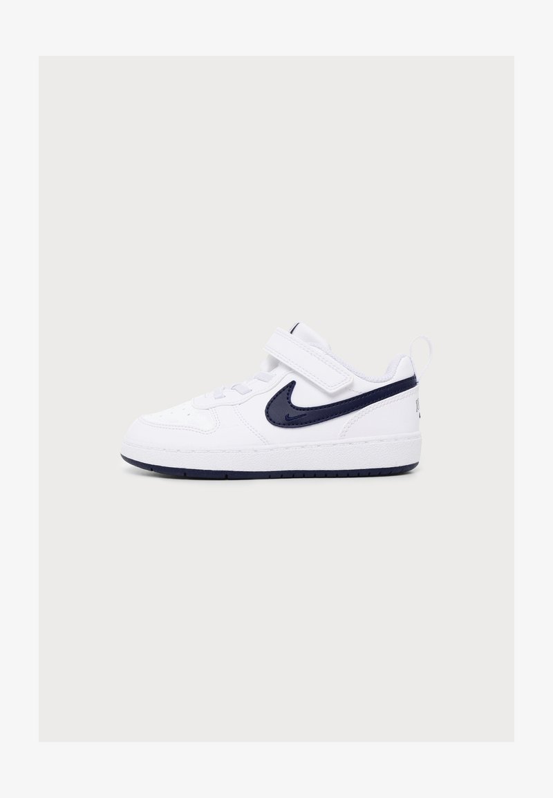 Λευκό αθλητικό παπούτσι με μαύρο λογότυπο Nike, ιμάντα βέλκρον και κορδόνια, εμφανίζεται από το πλάι σε ένα απλό φωτεινό φόντο.