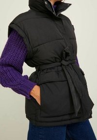 Černá prošívaná vesta se dvěma předními kapsami, páskem v pase; má vysoký límec a krátké rukávy. Nosená přes fialový pletený svetr.