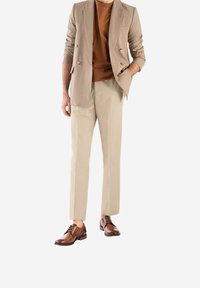 Beige dubbelzijdig blazer over een bruine shirt, gecombineerd met lichtbeige broeken en bruine leren schoenen, met veters en een strakke uitstraling.