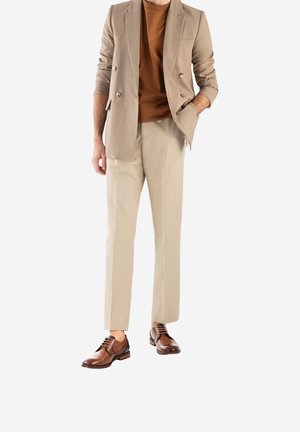 Blazer beige croisé sur une chemise marron, associé à un pantalon beige clair et des chaussures en cuir marron, avec lacets et un design élégant.