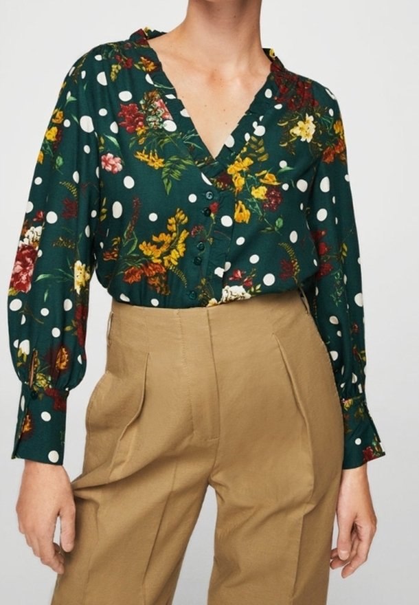 Femme portant un chemisier vert à pois blancs et motif floral, associé à un pantalon beige taille haute.