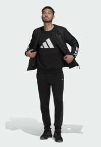Zwarte bomberjack met een gewatteerde textuur, zwarte sweatshirt met een wit logo, zwarte joggingbroek en zwarte sportschoenen met rode accenten.
