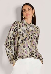Woman with straight dark hair wears gold statement earrings, a leopard print blouse with purple and beige tones, and white pants, hands in pockets.