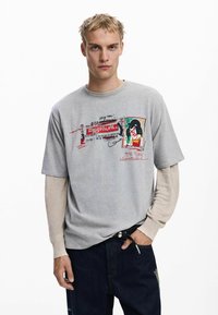 Šedé tričko s krátkym rukávom s grafickým potlačou a textom; vrstvené nad béžovým topom s dlhým rukávom; v kombinácii s tmavomodrými džínsami.