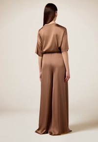 Combinaison en satin marron avec des manches courtes, design à jambes larges et texture lisse. La vue arrière met en évidence la coupe décontractée et la silhouette fluide.
