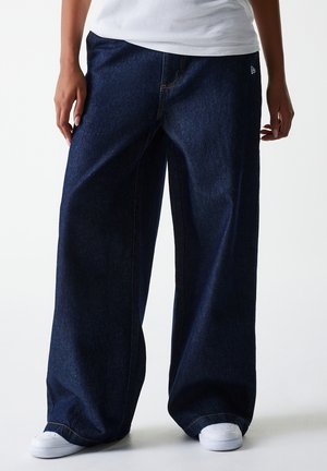 Dark blue wide-leg denim pants with visible stitching and a logo accent. Worn with white sneakers for casual styling.