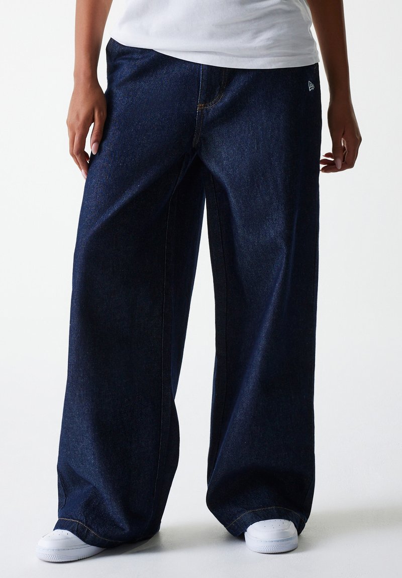 Pantalon en denim large bleu foncé avec coutures visibles et accent de logo. Porté avec des baskets blanches pour un style décontracté.