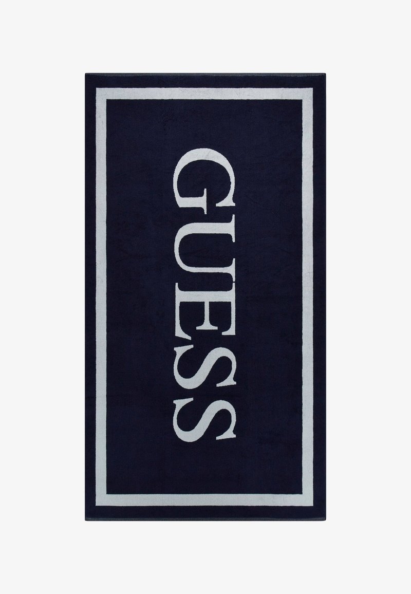 Marineblauw vloerkleed met een witte rechthoekige rand waarin het woord "GUESS" in grote, vetgedrukte witte letters over het midden staat. Zachte textuur.
